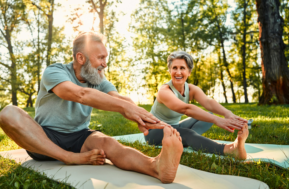 Flexible exercises for body. Sporty man and woman with grey hair stretching on yoga mats with hands to one leg during outdoors workout. Happy married couple with bare feet warming up together at park